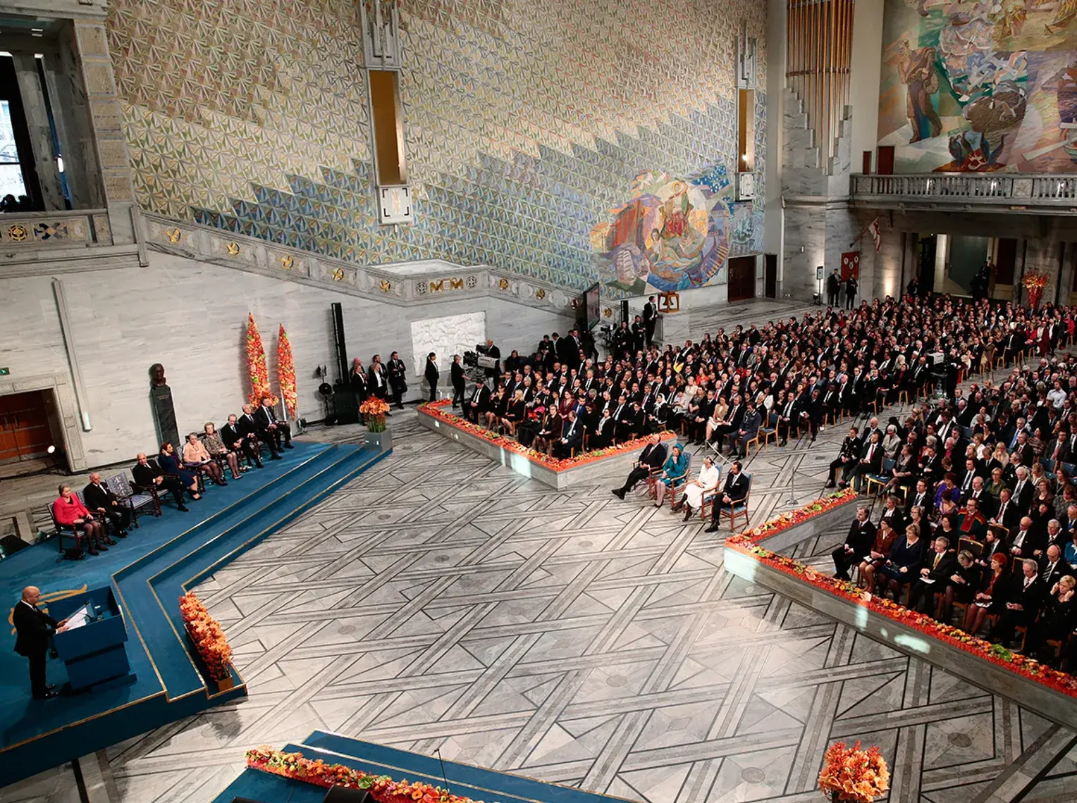 Government and Diplomacy — A formal award ceremony in a grand marble hall with murals, a speaker at the lectern and a large seated audience