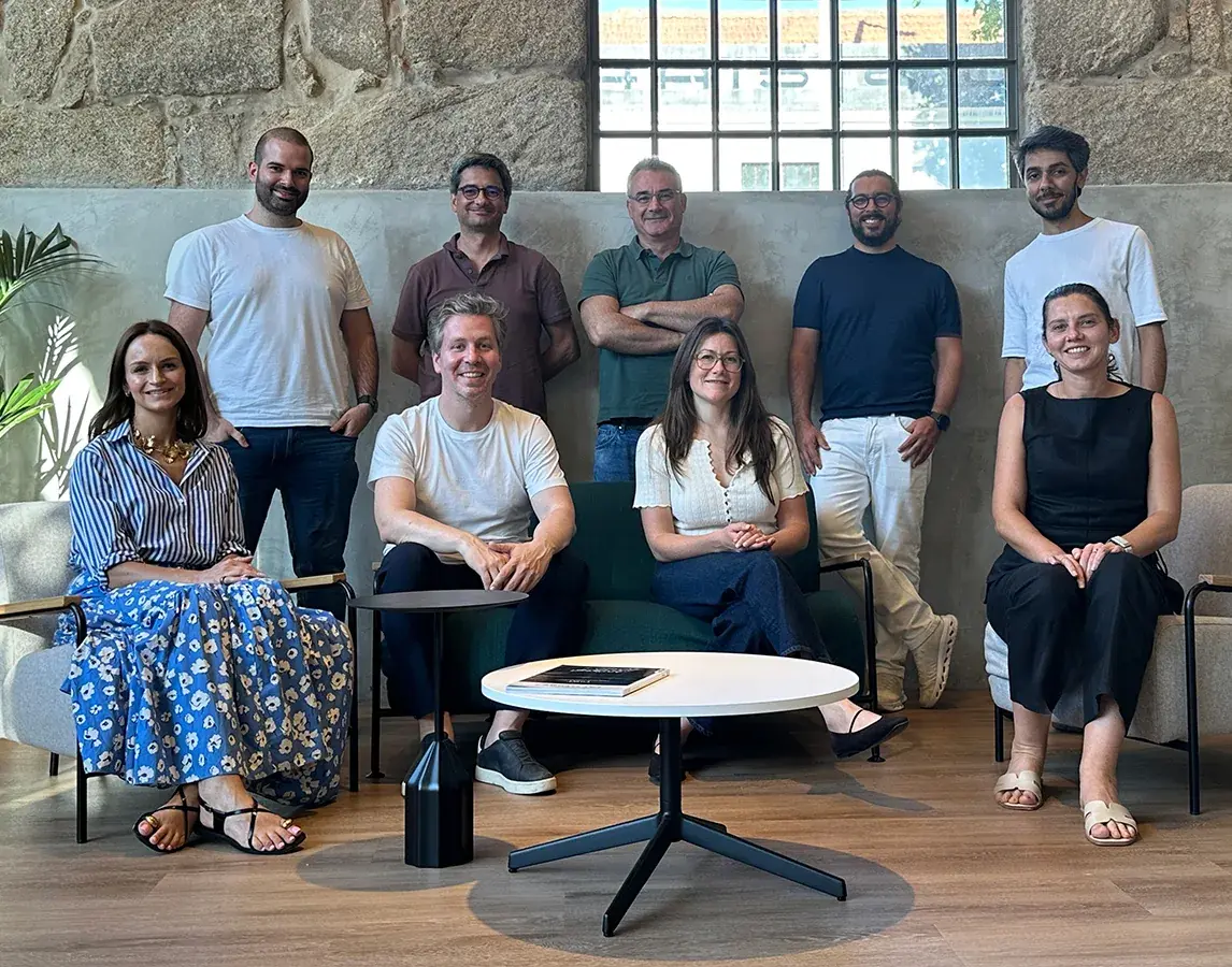 The zkipster leadership team, nine people smiling in a bright office with stone walls and a large window. Five stand at the back and four sit on sofas and armchairs around a round white coffee table.
